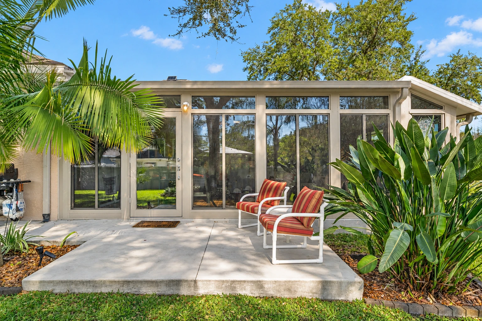 Enclosed patio room with broad glass doors and windows, flanked by tropical plants and two striped chairs.