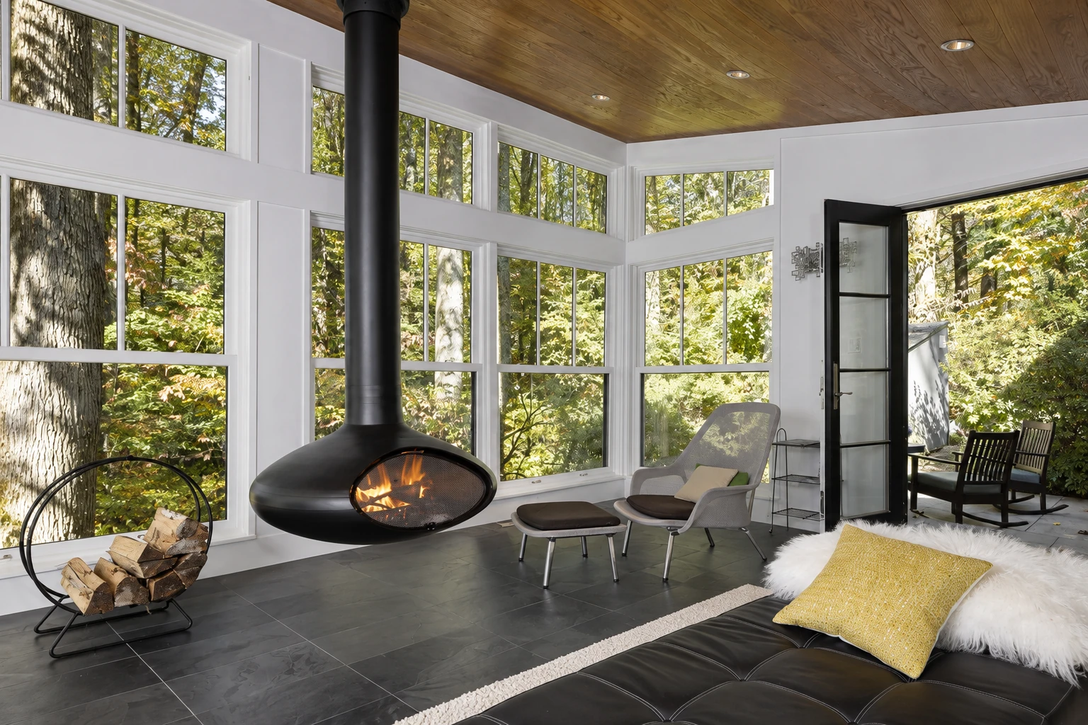 A bright enclosed patio room has tall windows, a wood ceiling, and a suspended fireplace.