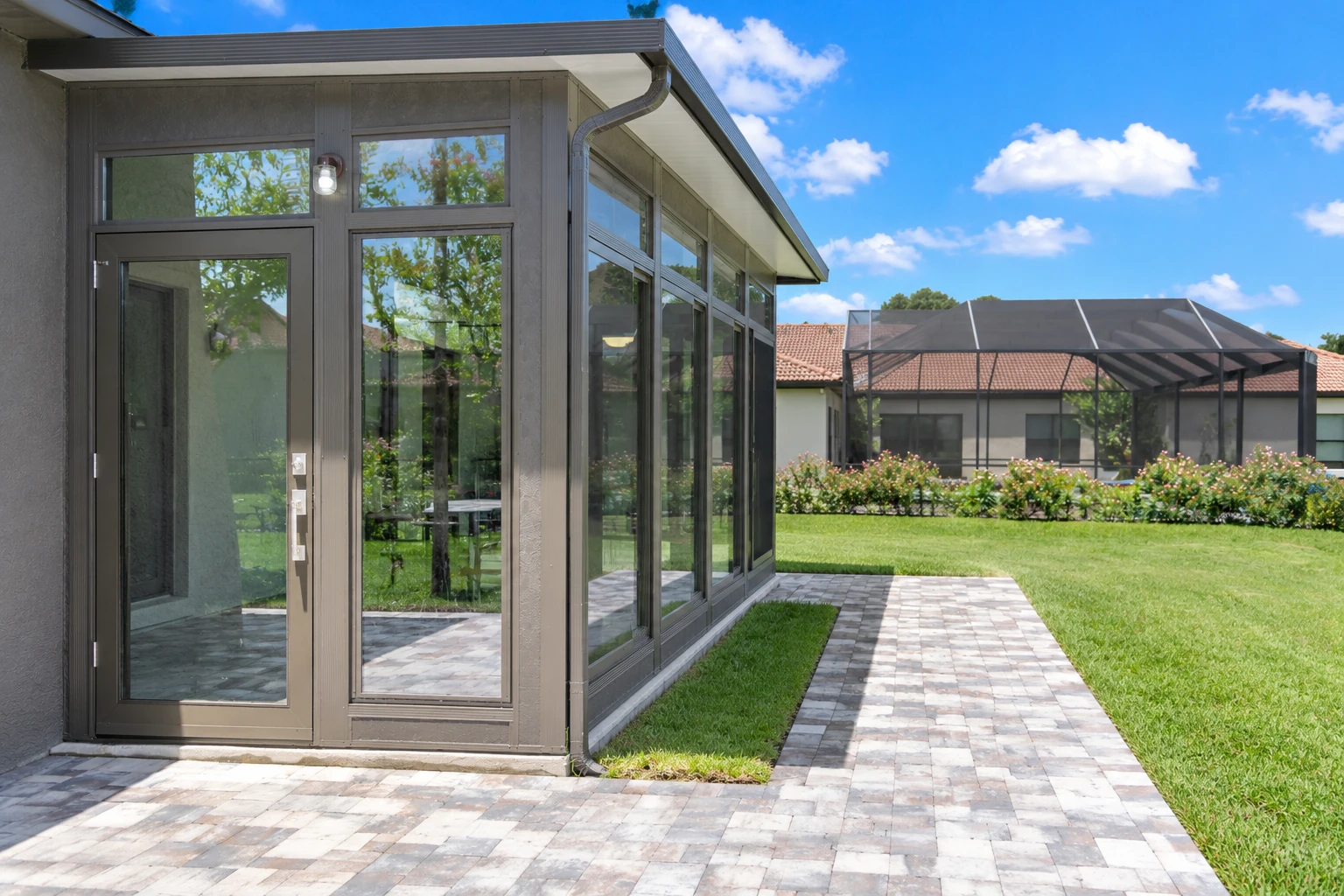 A gray-framed glass sunroom addition opens onto a paved patio beside a green lawn.