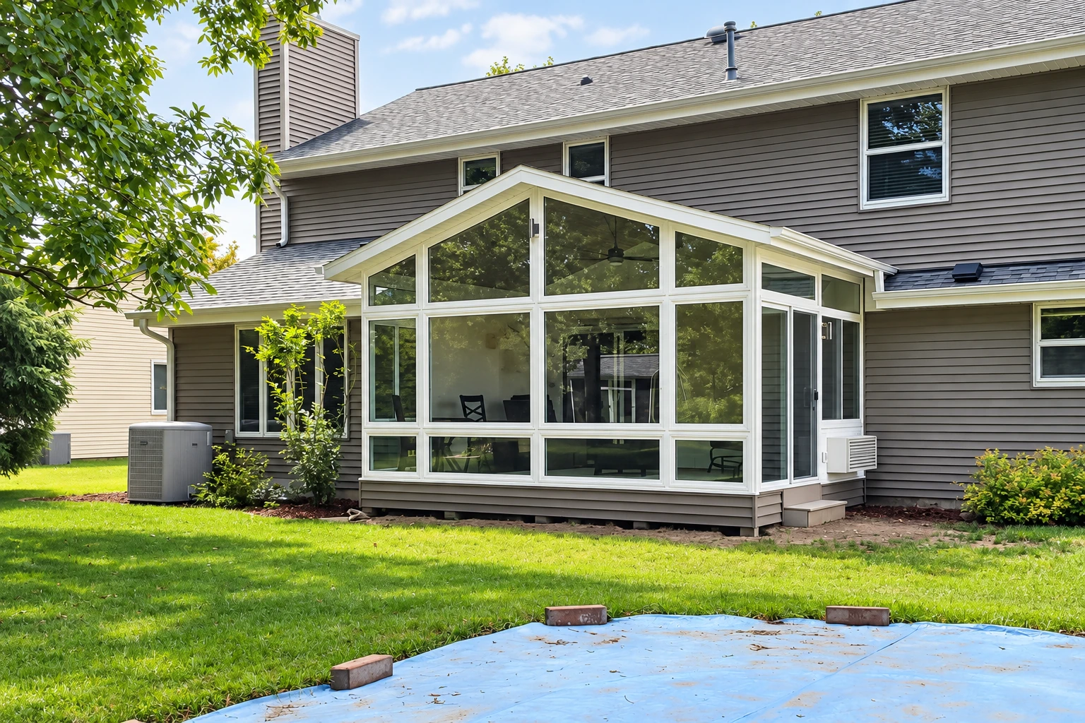 White-framed glass sunroom addition on a gray-sided house, with a peaked roof and lawn in front.