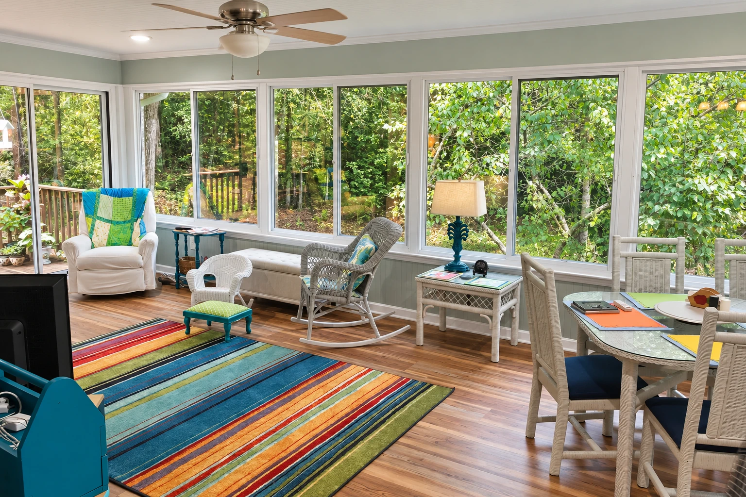 Bright sunroom with broad white-framed windows, ceiling fan, colorful striped rug, and seating and dining areas.